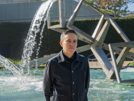 Mairead Sullivan wearing a black collared shirt and black jeans. She stands in front of Loyola Marymount University Hall's fountain that has a steel abstract sculpture in the middle of the fountain pool. The picture is taken in the morning, allowing the sun to glint off the falling water of the fountain and its pool behind Mairead Sullivan. She looks at the camera with a professional small smile.